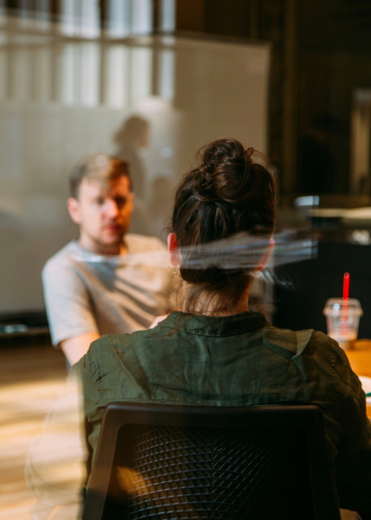 A client seen from behind speaking with a male therapist across a glass-walled consultation room