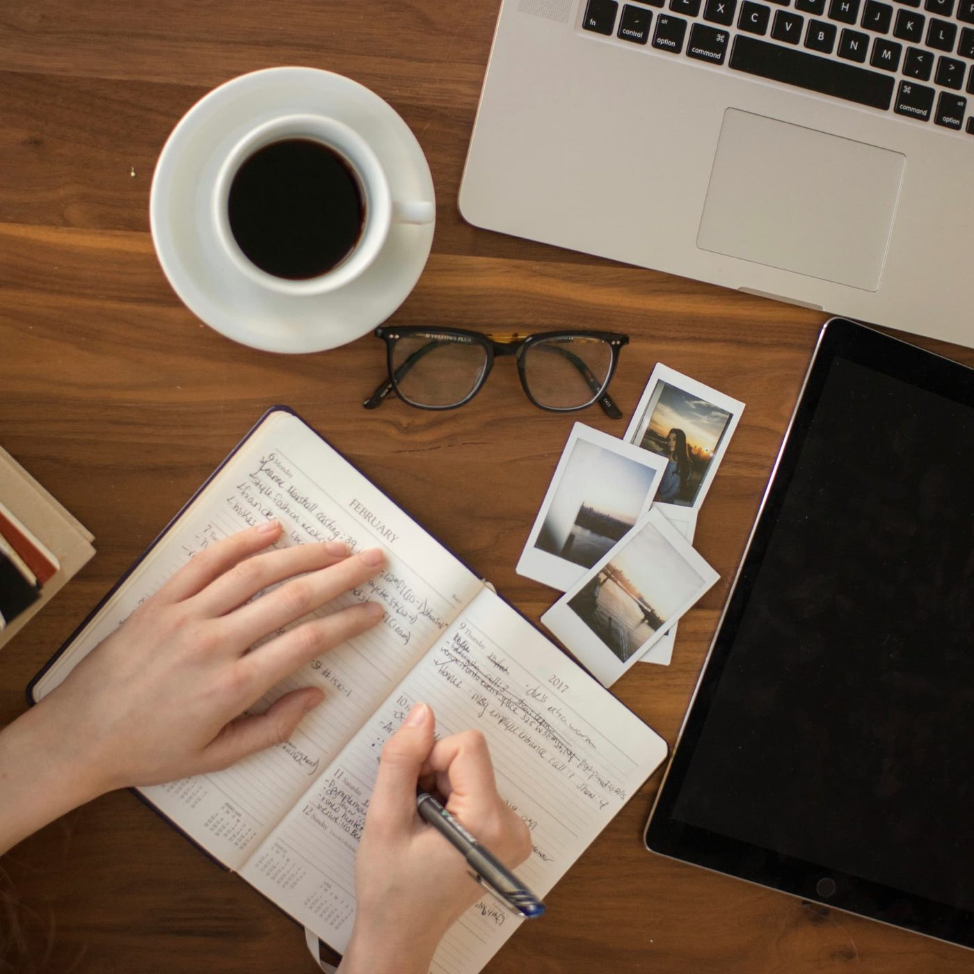 Hands writing in a diary on a wooden desk beside a coffee, glasses, a laptop and printed photographs