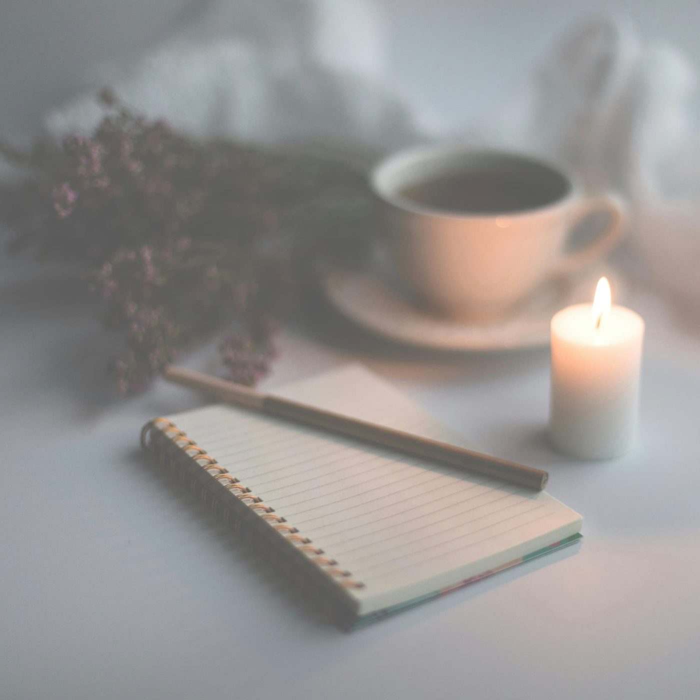 A small lined notepad and pen on a pale table beside a lit candle, a cup of tea and dried flowers