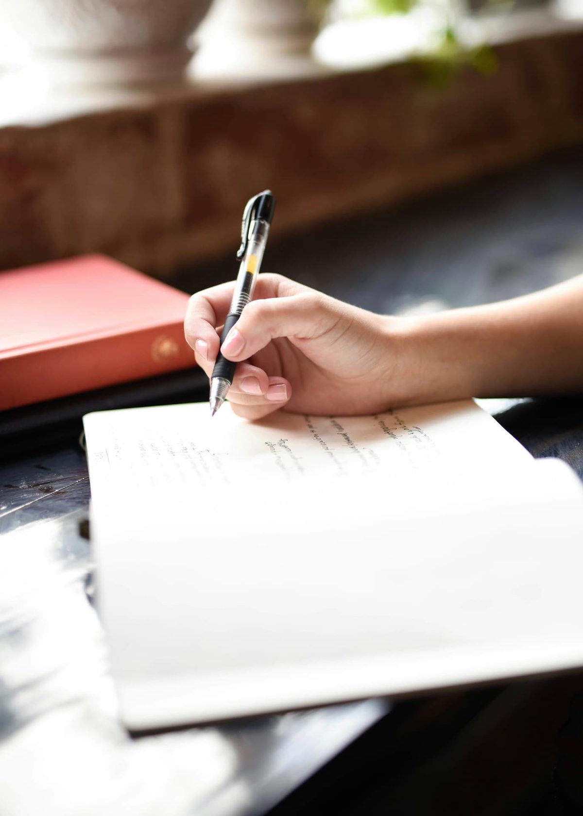 A hand holding a pen and writing in an open notebook on a dark table in warm window light