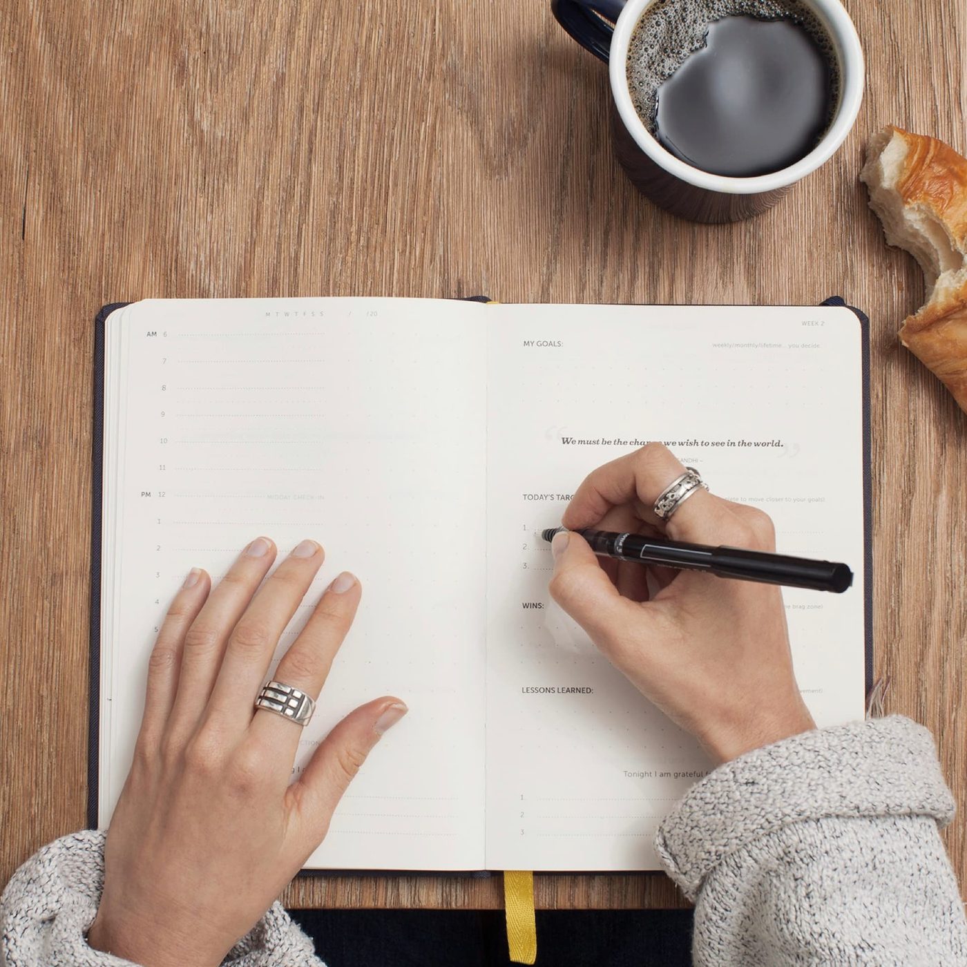 Overhead view of hands writing daily targets in a planner beside a black coffee and a croissant