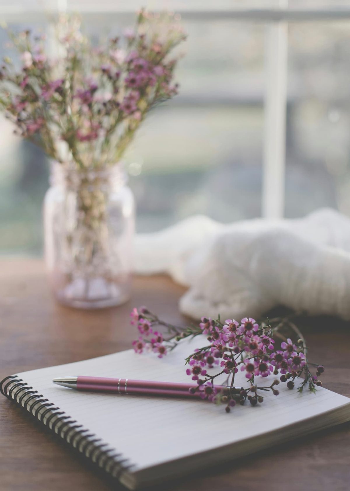 A spiral notebook and pink pen on a wooden table with sprigs of pink waxflower and a jar of blooms by a window