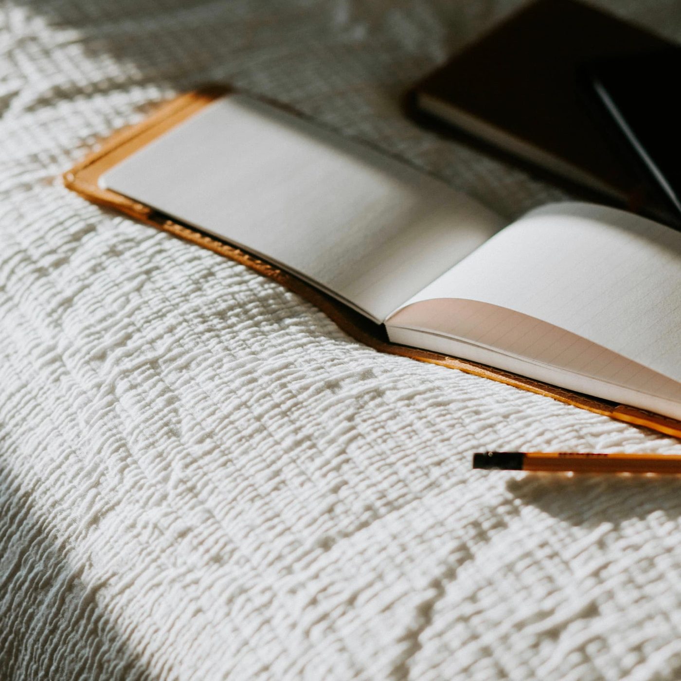 An open blank notebook in a tan leather cover resting on a textured white bedspread with a pencil beside it