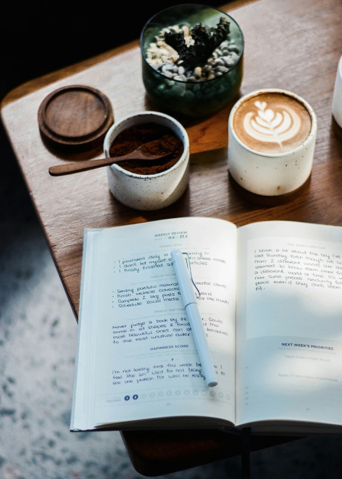 An open guided journal showing a handwritten weekly review beside a coffee on a wooden table