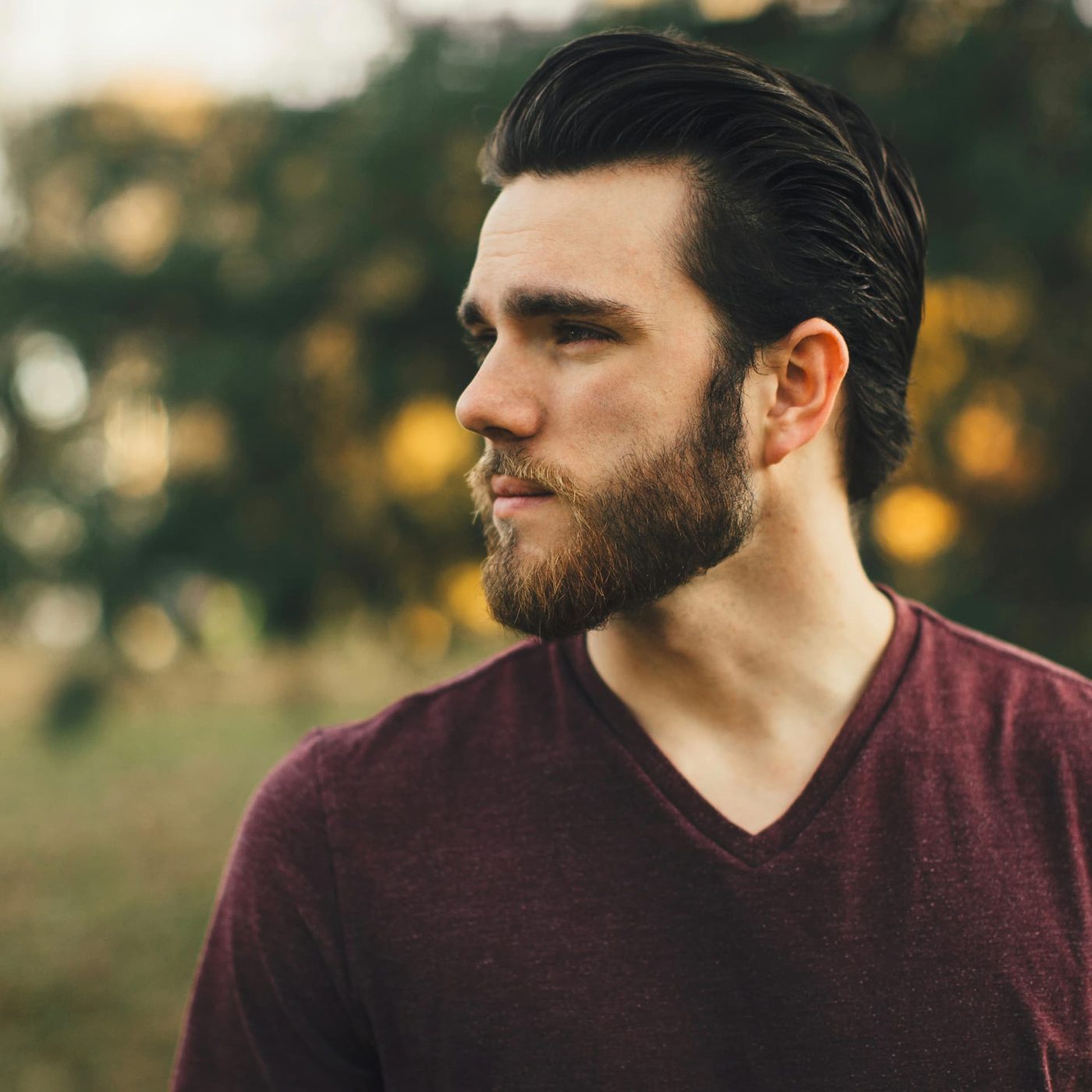 A bearded man in a burgundy t-shirt looking into the distance in a park at golden hour