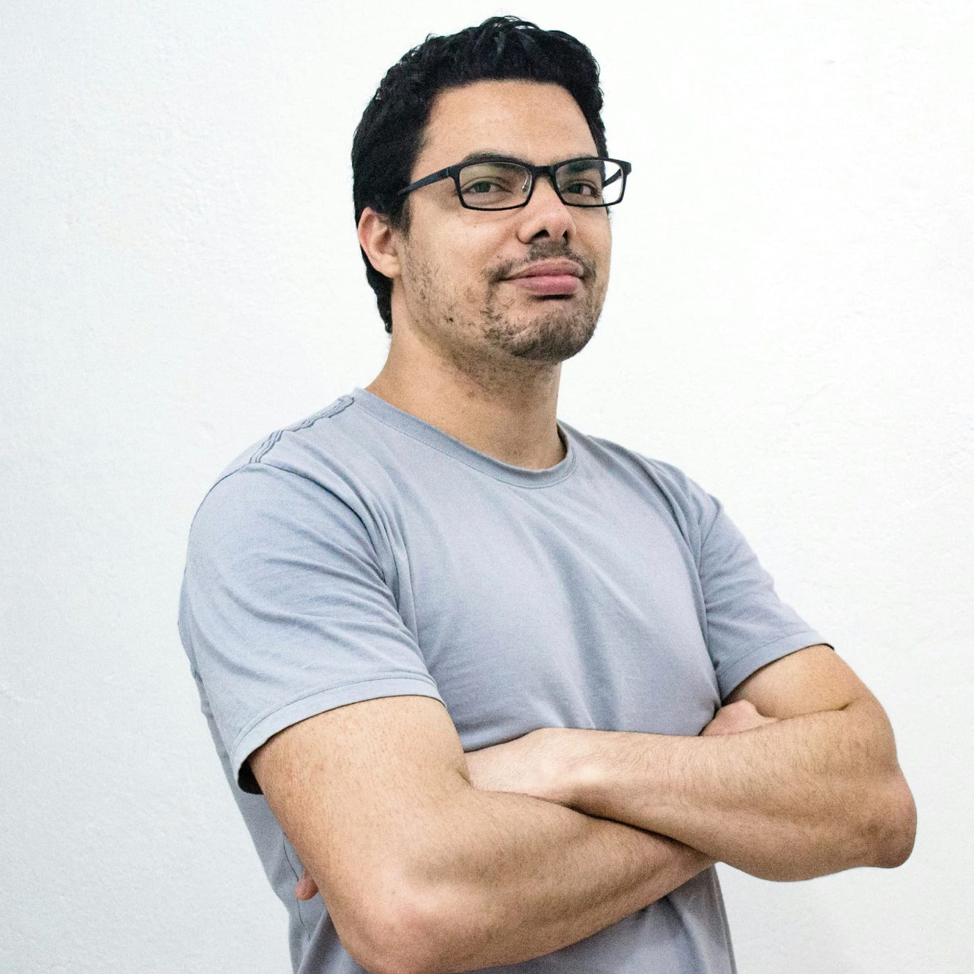 A man in a grey t-shirt and glasses standing with his arms folded against a plain white wall