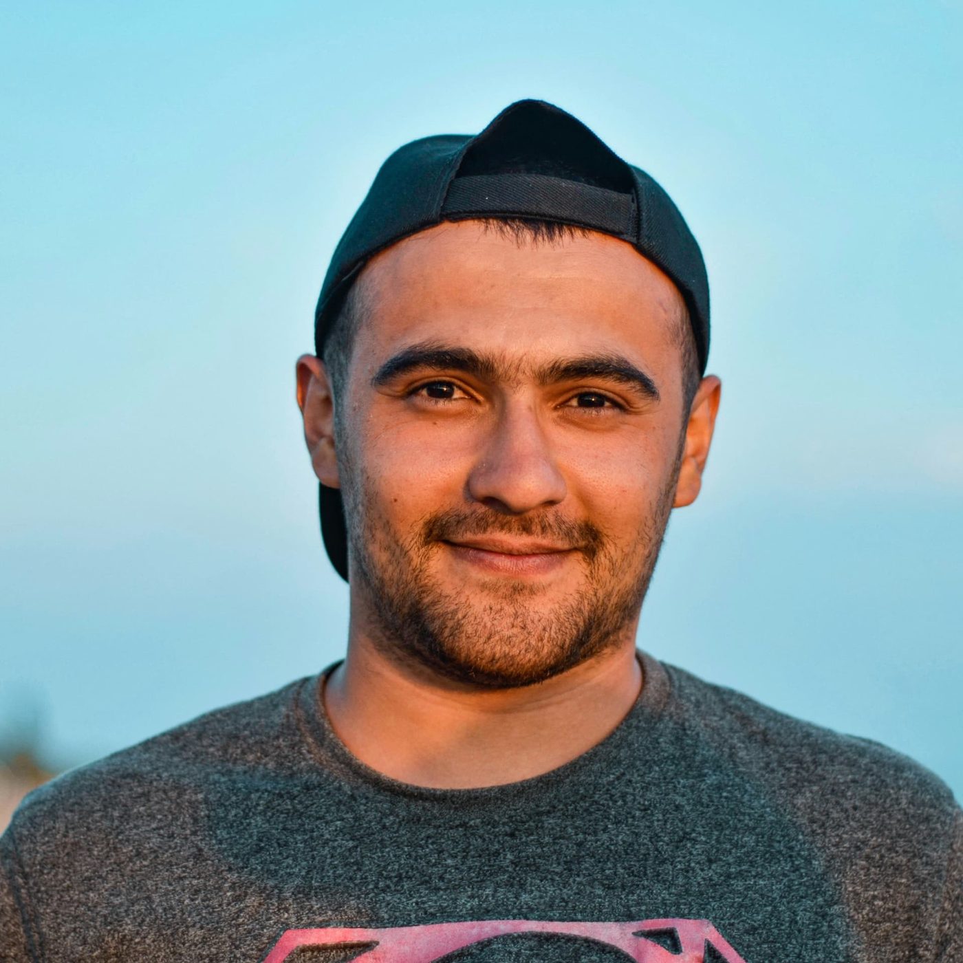 A man in a backwards black cap smiling gently against a pale blue evening sky