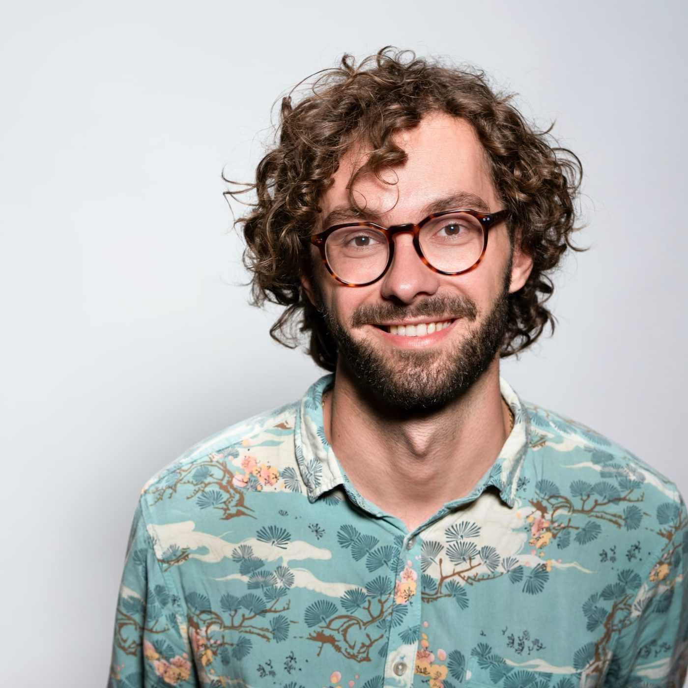 A man with curly hair, glasses and a patterned shirt smiling against a plain light background