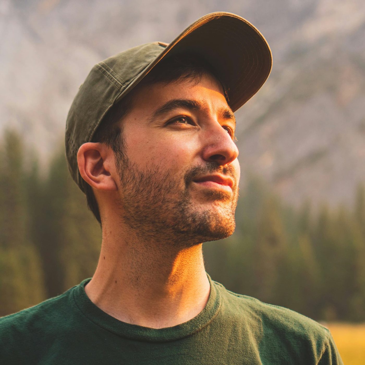 A man in a green cap looking upwards in warm evening light with pine trees and a mountainside behind him