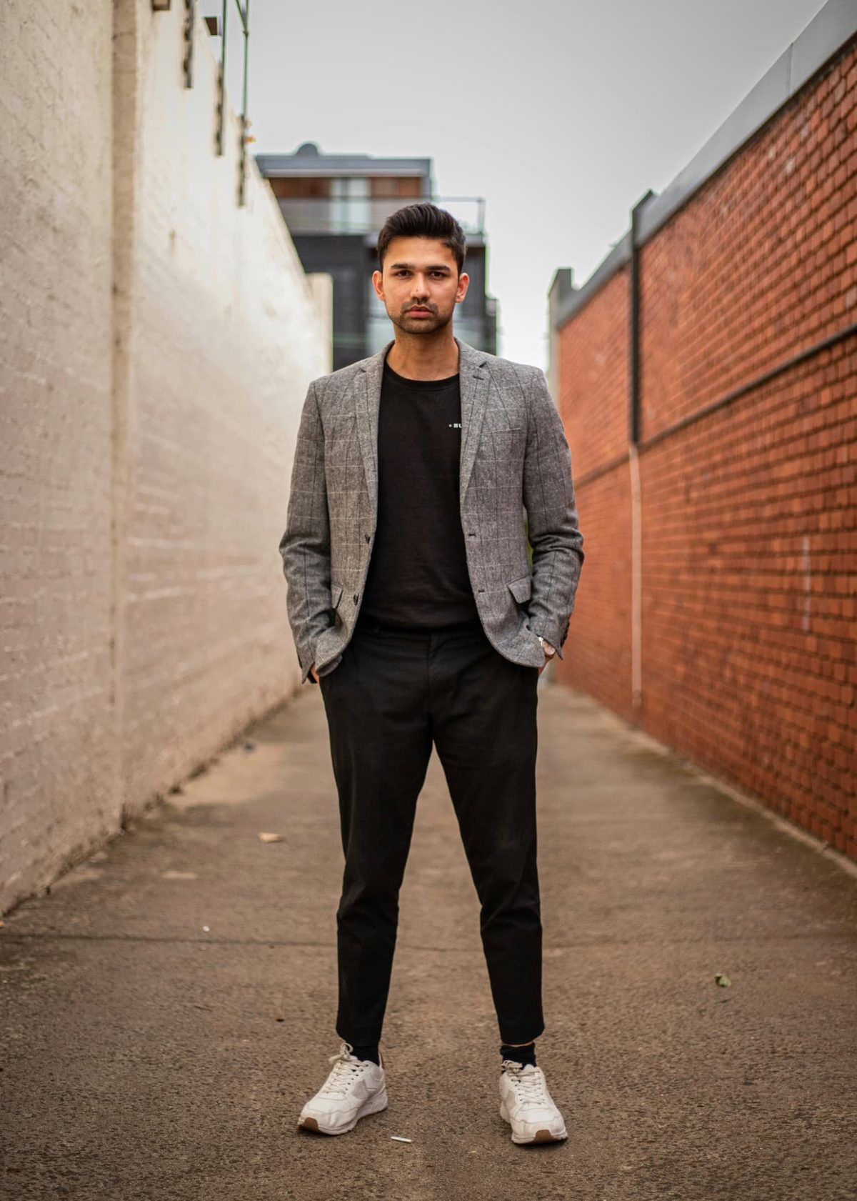 A man in a grey checked jacket and trainers standing with his hands in his pockets in a narrow city lane