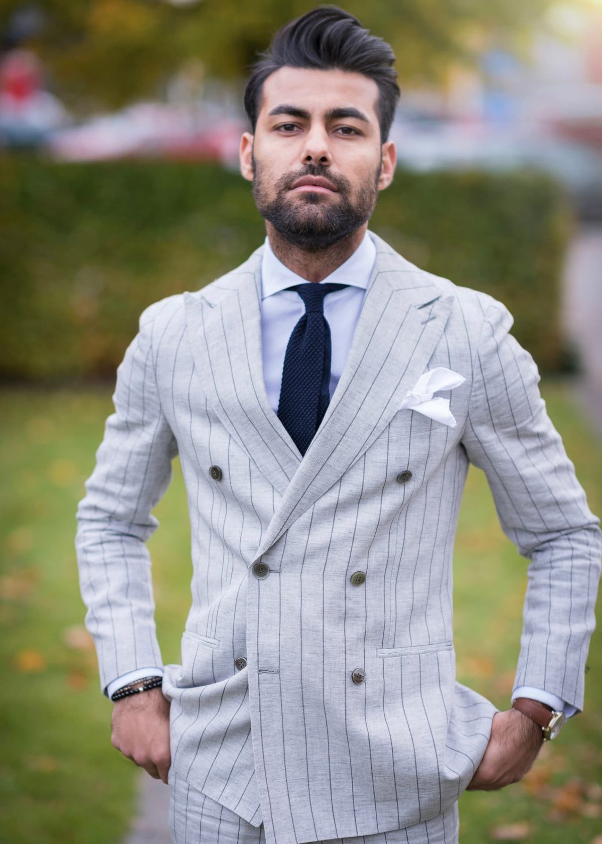 A bearded man in a light pinstripe suit standing in a park with autumn leaves behind him