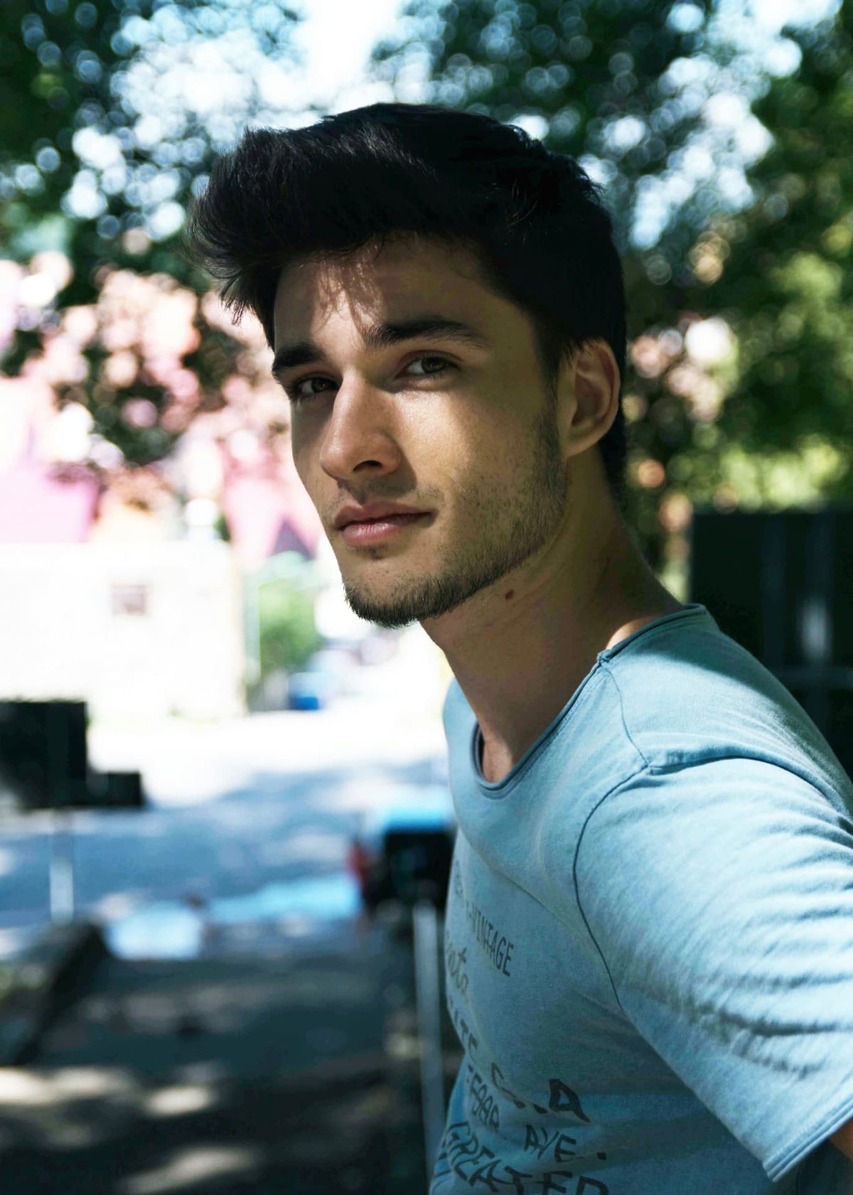 A young man in a pale t-shirt sitting outdoors in dappled summer shade, looking towards the camera