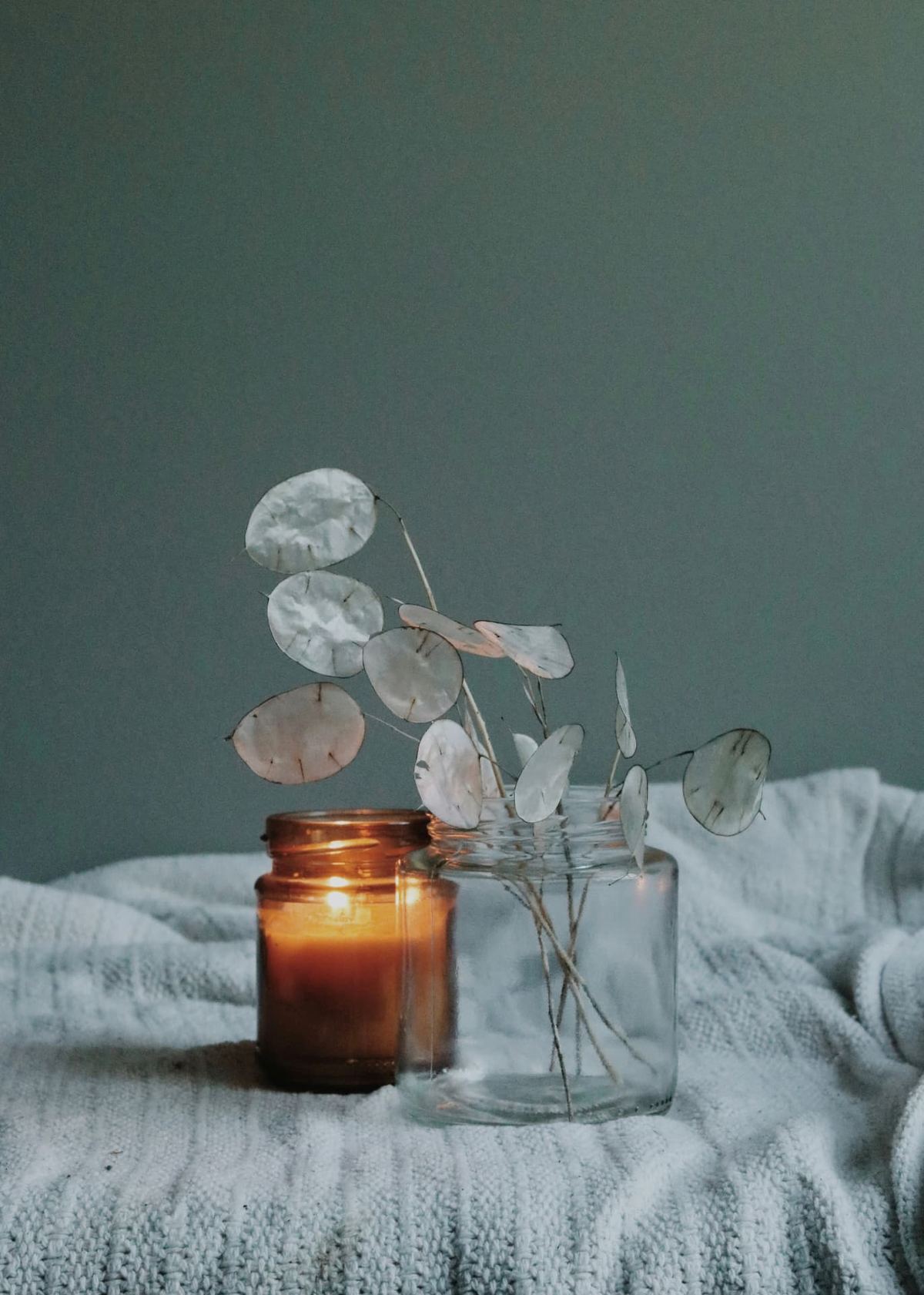 A lit candle in an amber jar beside dried honesty stems in a glass jar, on a soft knitted blanket