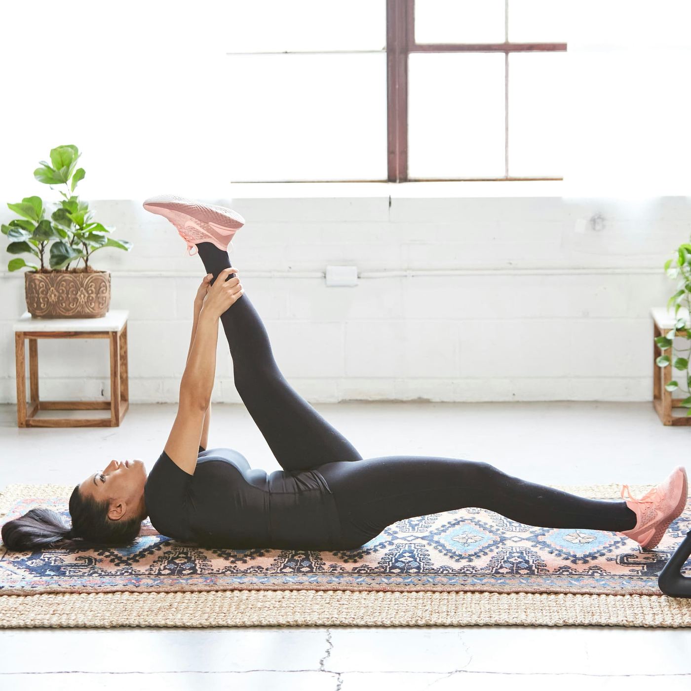 A woman lying on a rug in a bright room, holding one leg up in a gentle hamstring stretch