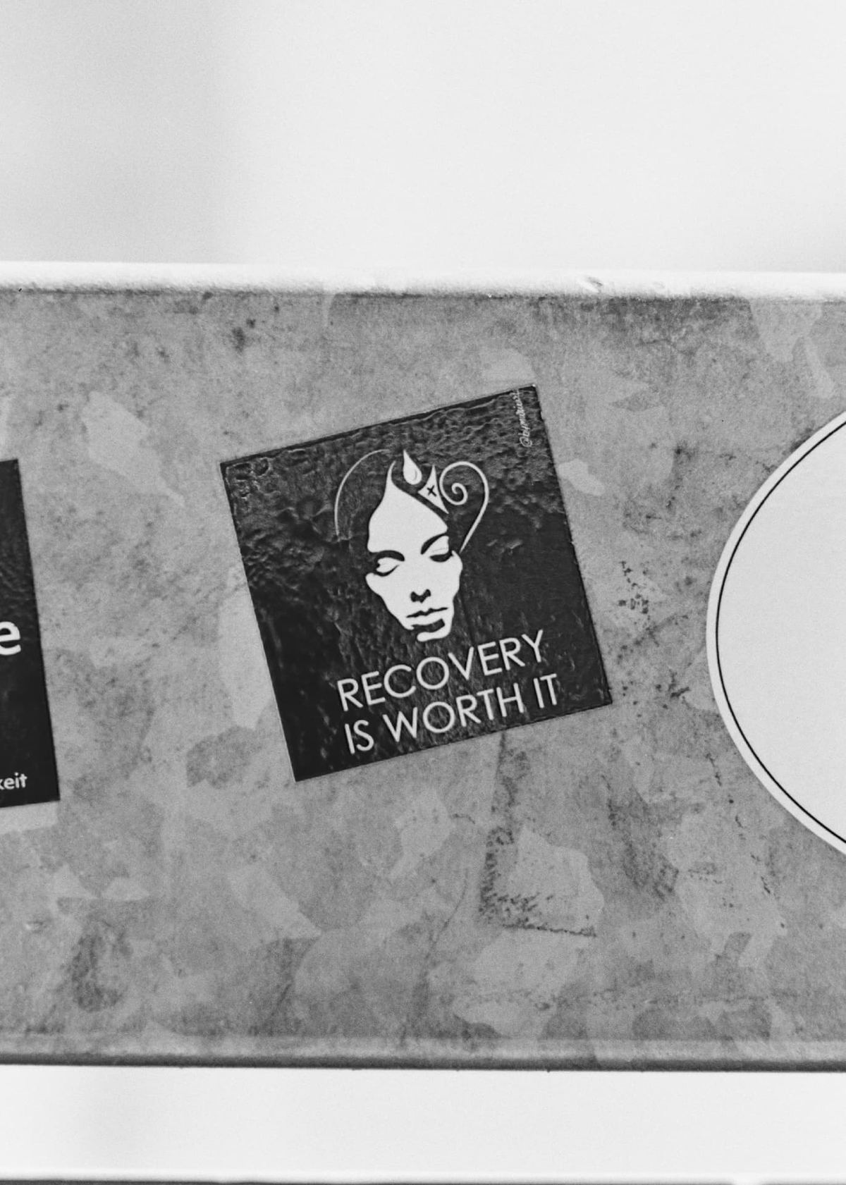 A black and white sticker on a metal surface reading 'Recovery is worth it' beside an illustrated calm face