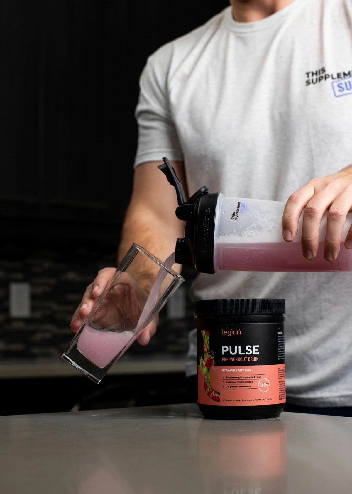 A person pouring a pink sports drink from a shaker bottle into a glass on a kitchen worktop