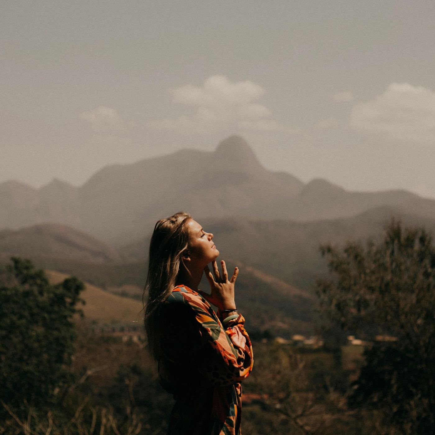 A woman standing on a hillside with her face turned up to the light, hazy mountains behind her