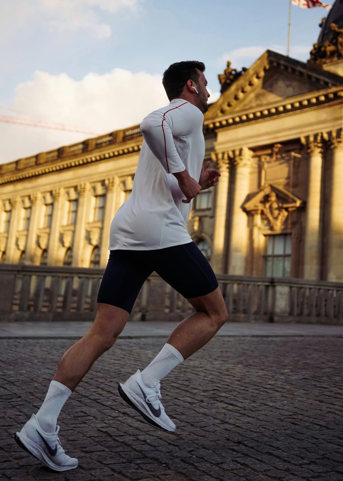 A runner in a white top running across cobblestones past a sunlit stone building in the early morning