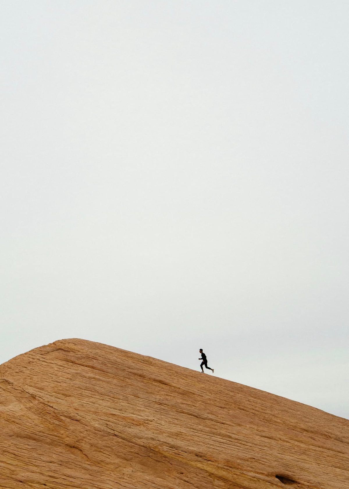 A small figure running along the crest of a wide sandstone ridge under a pale sky