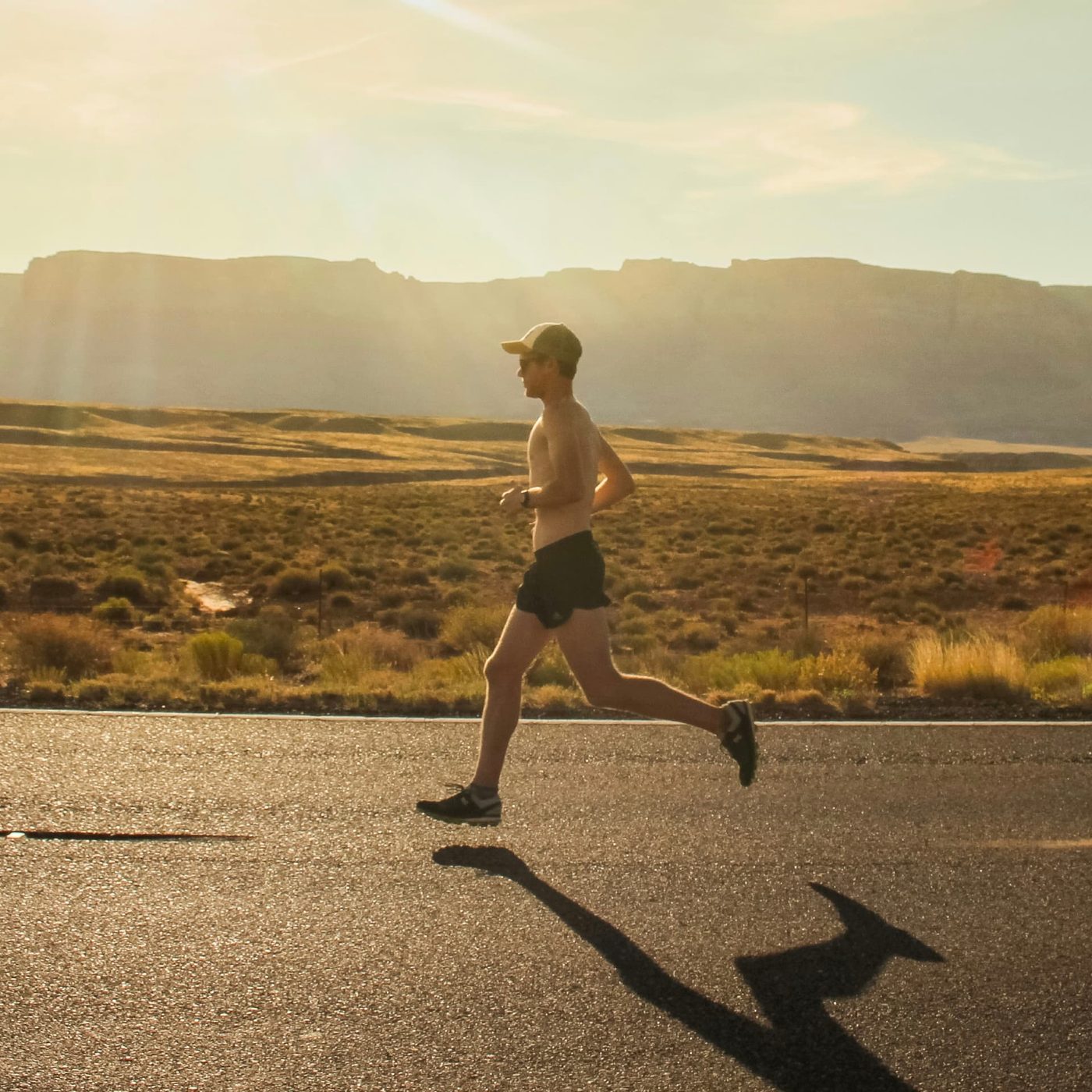 A runner in a cap moving steadily along an empty road at sunrise with open scrubland behind