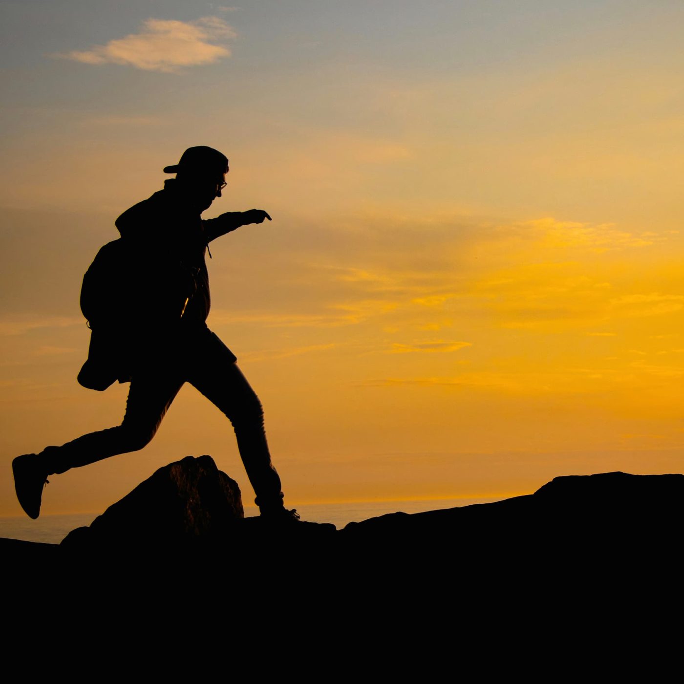 A person with a backpack silhouetted against an orange sunset, stepping across a gap in the rocks