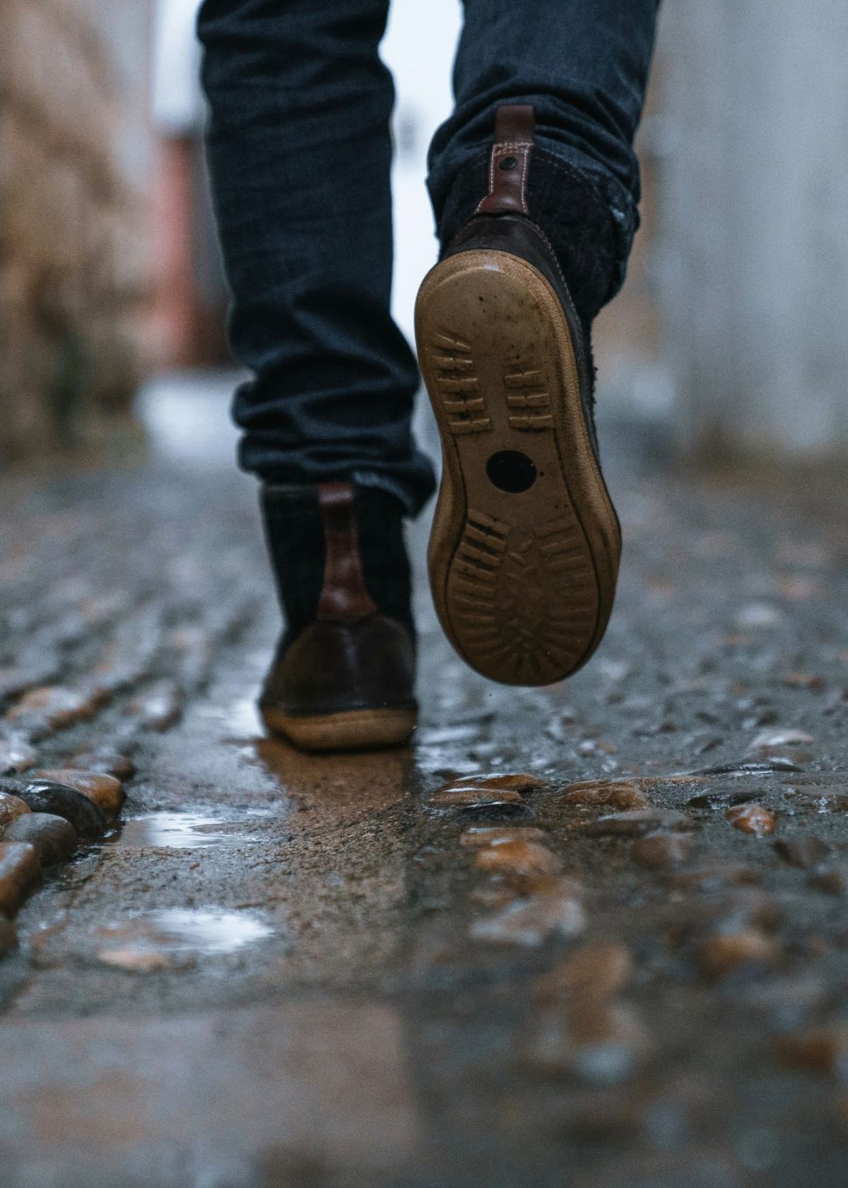 Close view of leather boots mid-step on wet cobbles, one sole lifted clear of a shallow puddle