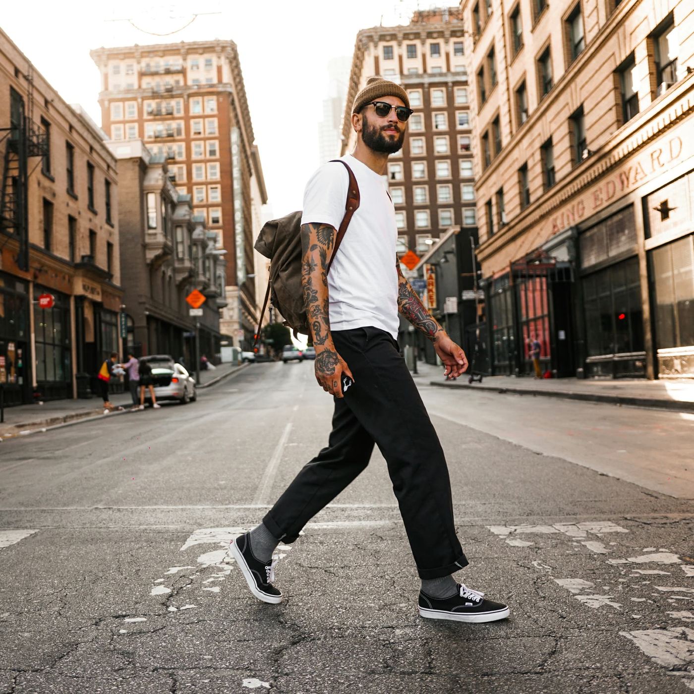 A man with a canvas rucksack crossing a wide city street between tall brick buildings in low sun