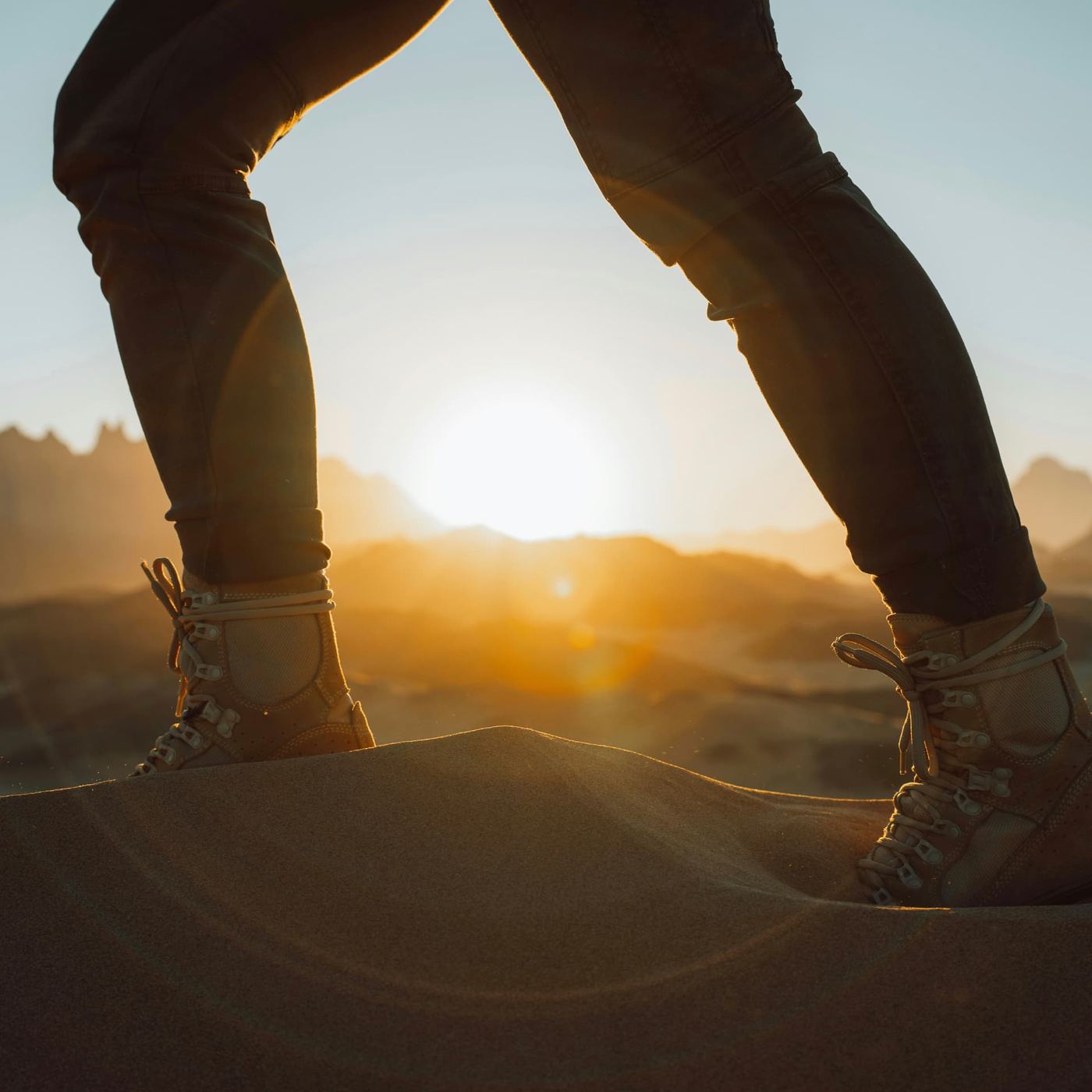 Walking boots planted on the crest of a sand dune with the sun setting behind distant hills