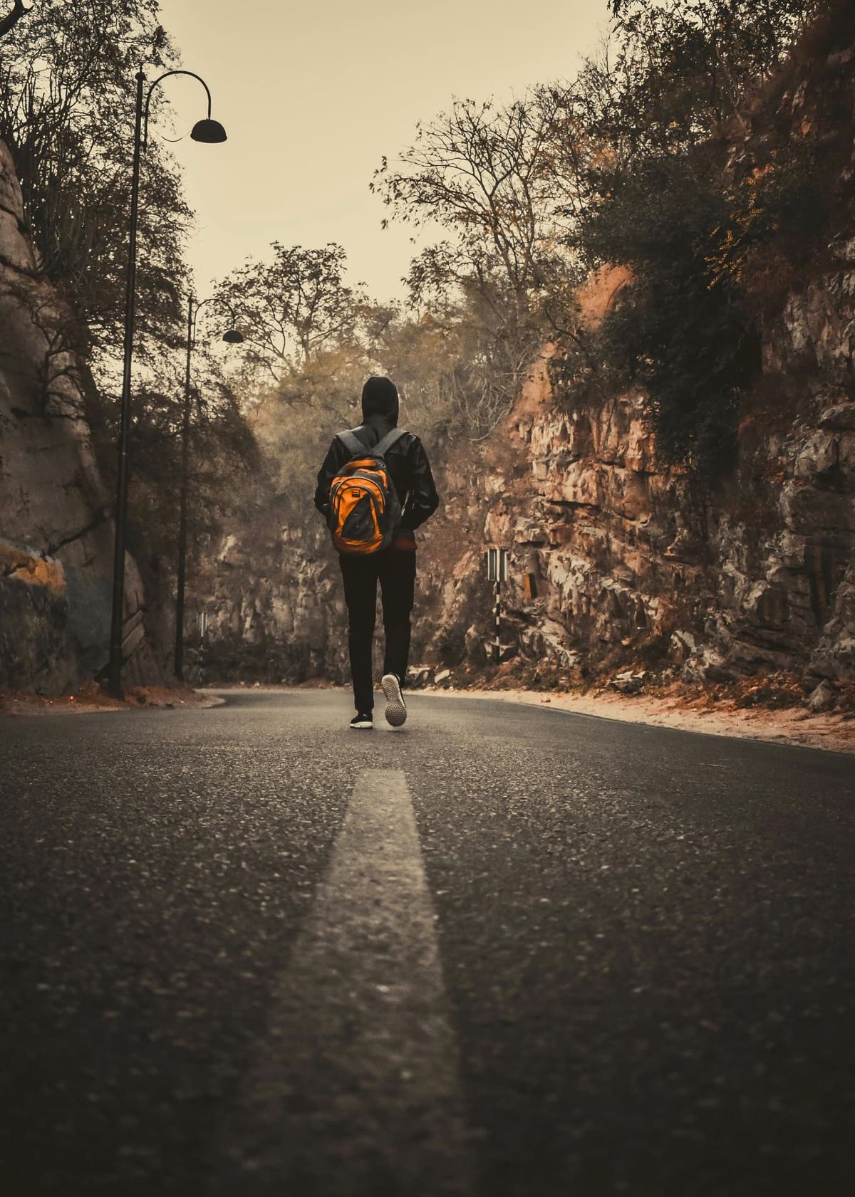 A lone walker in a hooded jacket heading up a quiet tree-lined road between rocky banks in autumn light
