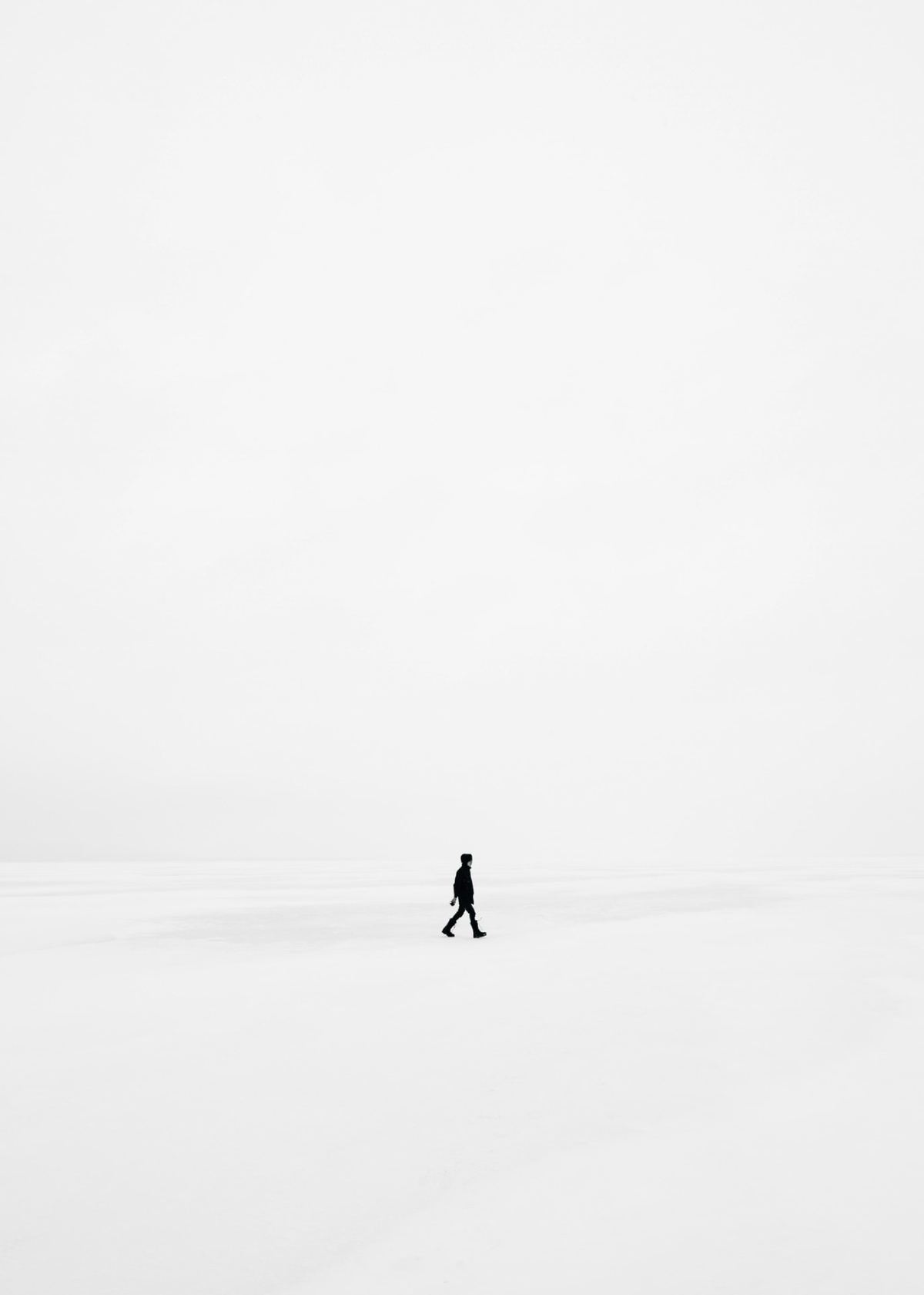 A single figure crossing a vast expanse of pale snow under an open white sky